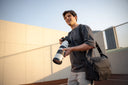 Outdoor photographer using Nomad sling bag while holding DSLR with telephoto lens. Secure compartments for gear.