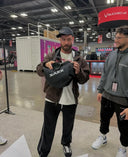 Person holding a Zakk helmet in a convention center setting with another person and a branded tent in the background.